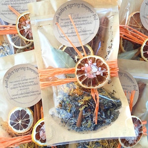 May include: A clear pouch labelled Aunt Ruth's Spring Simmer Pot containing dried herbs, lavender, and spices. The exterior is decorated with orange raffia, a dried orange slice, and pink salt crystals. Instructions for use are printed on the label.