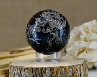 May include: A polished spherical stone featuring a deep dark blue base color with irregular, textured patches of metallic silver and grey mineral inclusions. The sphere is mounted on a small, clear circular acrylic stand for display.