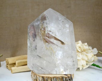 May include: A large, clear quartz crystal with brown and white inclusions. The crystal is shaped like a point and is sitting on a wooden base.