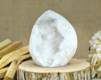 May include: A white quartz geode with a hollow center filled with crystalline structures. The geode has a smooth, rounded exterior and is positioned upright, displaying a jagged, sparkling interior cavity with translucent white mineral formations.