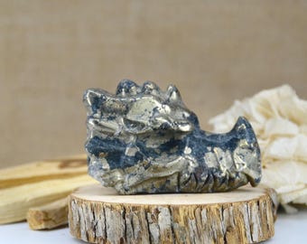 May include: A carved pyrite dragon head figurine featuring a metallic gold and dark gray mottled surface with sharp, angular details. The sculpture is positioned on a circular wooden slice base with visible bark edges and a natural wood grain texture.