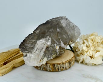 May include: A large, smoky quartz crystal with a clear, faceted point. The crystal is resting on a wooden slice and is surrounded by a white background.
