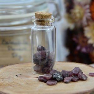 May include: A small glass bottle with a cork stopper filled with small, dark purple gemstones. The bottle is sitting on a wooden surface with more gemstones scattered around it.