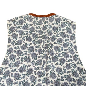 May include: A sleeveless garment featuring a cream-colored paisley pattern with green and purple accents. The neckline is trimmed with a brown corduroy fabric. The garment is likely a top or dress, showcasing a vintage or bohemian style.