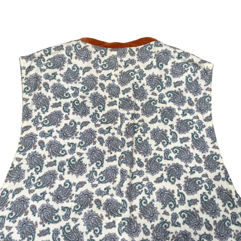 May include: A sleeveless garment featuring a cream-colored paisley pattern with green and purple accents. The neckline is trimmed with a brown corduroy fabric. The garment is likely a top or dress, showcasing a vintage or bohemian style.