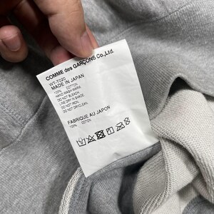 K&ouml;nnte beinhalten: Nahaufnahme eines grauen Sweatshirts mit Etikett. Auf dem Etikett steht "COMME des GAR&Ccedil;ONS Co.,Ltd." und "MADE IN JAPAN". Auf dem Etikett sind auch Waschanweisungen abgedruckt. Das Sweatshirt besteht aus 100 % Baumwolle.
