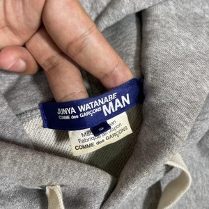K&ouml;nnte beinhalten: Nahaufnahme eines grauen Hoodies mit wei&szlig;em Kordelzug. Ein blaues Etikett lautet "JUNYA WATANABE COMME des GAR&Ccedil;ONS MAN". Darunter steht auf einem wei&szlig;en Etikett "Made in Japan" und "Fabriqu&eacute; au Japon COMME des GAR&Ccedil;ONS".