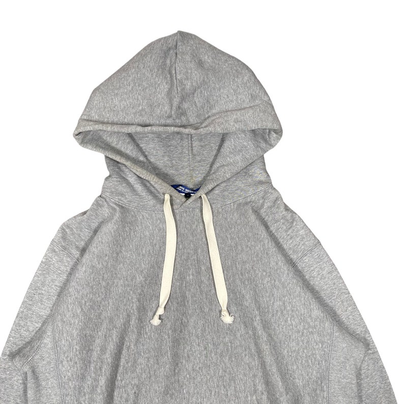K&ouml;nnte beinhalten: Ein melierter grauer Kapuzenpullover mit Kapuze und Kordelzug sowie wei&szlig;en Kordeln. Das Sweatshirt hat eine K&auml;ngurutasche. Der Stoff scheint ein weiches, bequemes Material zu sein, das sich f&uuml;r Freizeitkleidung eignet. Der Hoodie ist ein klassisches, vielseitiges Kleidungsst&uuml;ck.