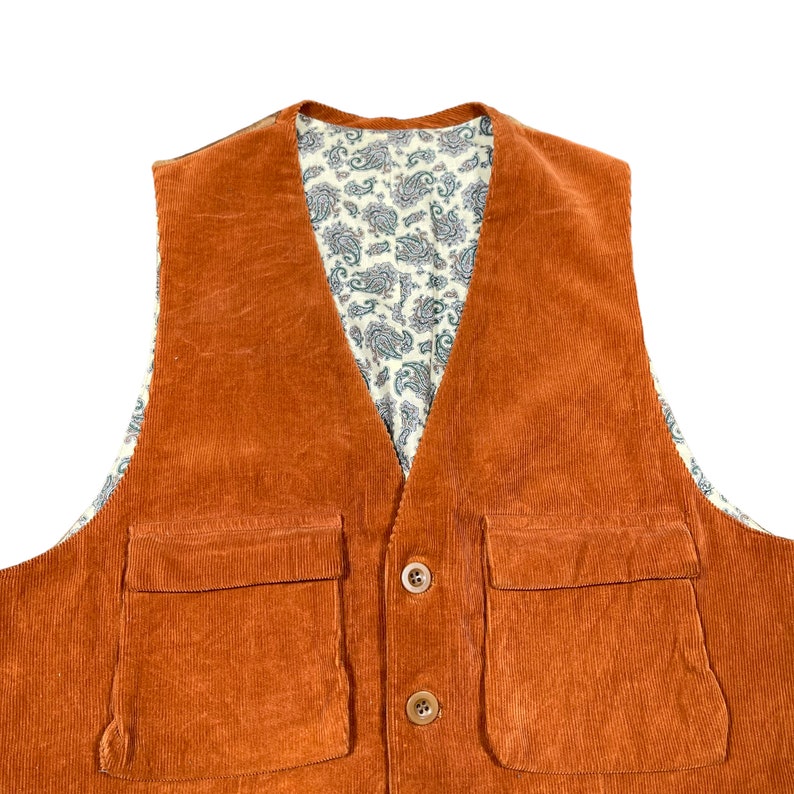 May include: An orange corduroy vest with two front pockets and two brown buttons. The interior lining features a cream-colored paisley pattern with blue and green accents. The vest is a classic style.