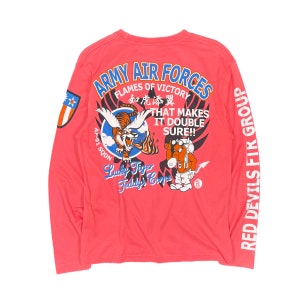 Può includere: Una maglietta a maniche lunghe color corallo con disegni grafici. Il retro presenta una tigre, un personaggio dei cartoni animati e il testo "ARMY AIR FORCES" e "Lucky Tiger Teddy's Corps". La manica sinistra ha una toppa. "Red Devils FIR Group" è stampato sulla manica destra.