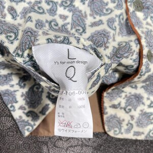 May include: Close-up of a cream-colored shirt with a paisley pattern in shades of gray, green, and purple. The shirt has a brown trim and a tag that reads "Y's for men design" with the letter "L" and the letter "Q".