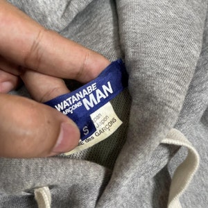 K&ouml;nnte beinhalten: Nahaufnahme eines grauen Hoodies mit wei&szlig;em Kordelzug. Ein blaues Etikett mit wei&szlig;em Text lautet "WATANABE MAN" und "COMME des GARCONS". Ein kleineres Etikett darunter lautet "Japan" und "COMME des GARCONS".
