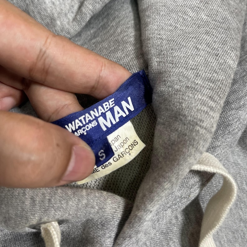 K&ouml;nnte beinhalten: Nahaufnahme eines grauen Hoodies mit wei&szlig;em Kordelzug. Ein blaues Etikett mit wei&szlig;em Text lautet "WATANABE MAN" und "COMME des GARCONS". Ein kleineres Etikett darunter lautet "Japan" und "COMME des GARCONS".