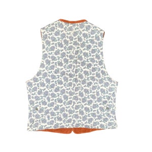 May include: A reversible vest with a paisley pattern on a cream background. The vest has a rust-colored trim and lining. The paisley pattern features blue and gray details. The vest is a classic style, suitable for layering.