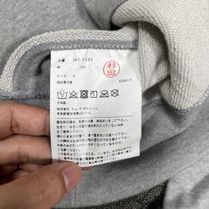 K&ouml;nnte beinhalten: Nahaufnahme eines grauen Sweatshirt-Etiketts mit japanischem Text und Pflegesymbolen. Das Etikett enth&auml;lt den Produktcode WT-T020, die Gr&ouml;&szlig;e S und den Text "100%" und "AD2017". Das Etikett ist an einem grauen Sweatshirt befestigt.