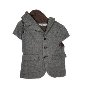 May include: A grey tweed blazer with short sleeves and a distinctive cut-out design. The blazer has a notched lapel, three black buttons, and two front pockets. The label reads "JUNYA WATANABE COMME des GARÇONS".
