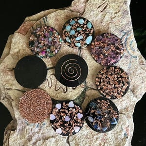 May include: A collection of handcrafted, circular pendants with various designs. Some pendants feature colorful stones and copper accents, while others have a solid black finish or a copper spiral. The pendants are displayed on a textured, light-colored surface.