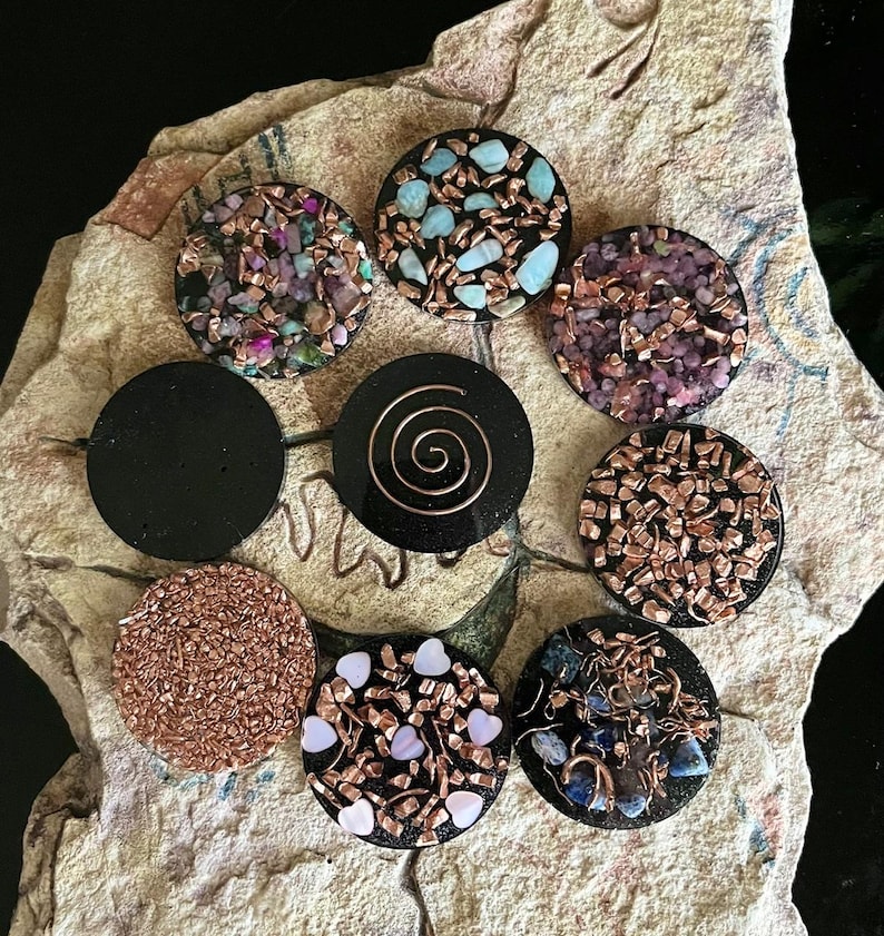 May include: A collection of handcrafted, circular pendants with various designs. Some pendants feature colorful stones and copper accents, while others have a solid black finish or a copper spiral. The pendants are displayed on a textured, light-colored surface.