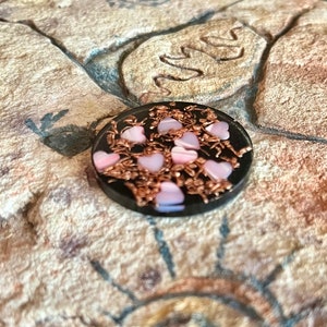 May include: A small, round, black object with pink heart-shaped accents and copper-colored wire detailing. The hearts are a soft pink color. The object is set against a textured, tan-colored surface.
