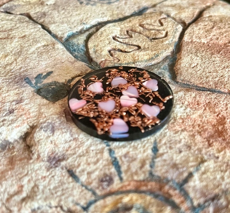 May include: A small, round, black object with pink heart-shaped accents and copper-colored wire detailing. The hearts are a soft pink color. The object is set against a textured, tan-colored surface.