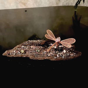 May include: A decorative sculpture featuring a copper wire dragonfly with mesh wings and a clear crystal body, mounted on a piece of dark driftwood. The wood surface is embedded with small, multicolored mineral stones and metallic fragments.