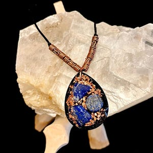 May include: A teardrop-shaped pendant necklace featuring deep blue lapis lazuli stones embedded in a black resin base with copper flakes. The pendant hangs from a black cord accented with two cylindrical copper beads with ribbed detailing.