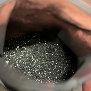 May include: A close-up view of a clear plastic bag filled with a dark, granular substance. The substance appears to be small, shiny particles, possibly a powder or fine granules. The bag is held open, revealing the contents.