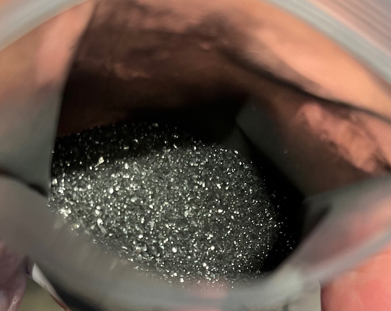 May include: A close-up view of a clear plastic bag filled with a dark, granular substance. The substance appears to be small, shiny particles, possibly a powder or fine granules. The bag is held open, revealing the contents.