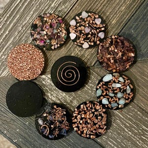 May include: A collection of eight round, black orgone energy devices. Each device features a unique design, including copper wire, crystals, and a spiral. The crystals are various colors, including pink, blue, and brown. The devices are arranged on a wooden surface.
