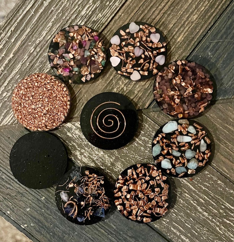 May include: A collection of eight round, black orgone energy devices. Each device features a unique design, including copper wire, crystals, and a spiral. The crystals are various colors, including pink, blue, and brown. The devices are arranged on a wooden surface.