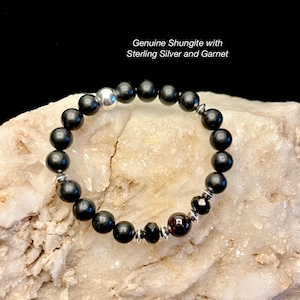 May include: A beaded bracelet featuring black, round stones, sterling silver accents, and a deep red garnet stone. The text "Genuine Shungite with Sterling Silver and Garnet" is displayed in the image.