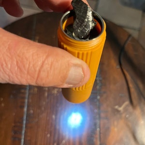 May include: An orange, cylindrical container with a silver interior holds a dark, textured object. The container is held by a hand, with the object partially visible. The background is a wooden surface.