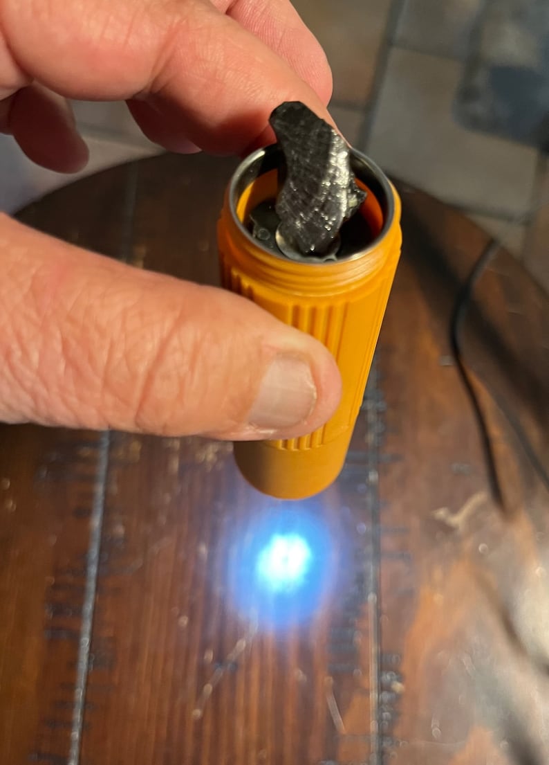 May include: An orange, cylindrical container with a silver interior holds a dark, textured object. The container is held by a hand, with the object partially visible. The background is a wooden surface.
