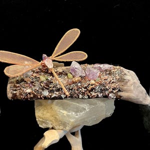 May include: A decorative piece featuring a copper wire dragonfly with mesh wings perched on a piece of driftwood. The wood is adorned with small, colorful gemstone chips, including purple amethyst, and rests on a translucent white crystal base.