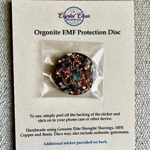 May include: An Orgonite EMF Protection Disc, featuring a mix of copper, resin, and gemstones, is displayed in a clear plastic package. The disc is round with a dark, speckled appearance. The packaging includes text about the product's use and materials.