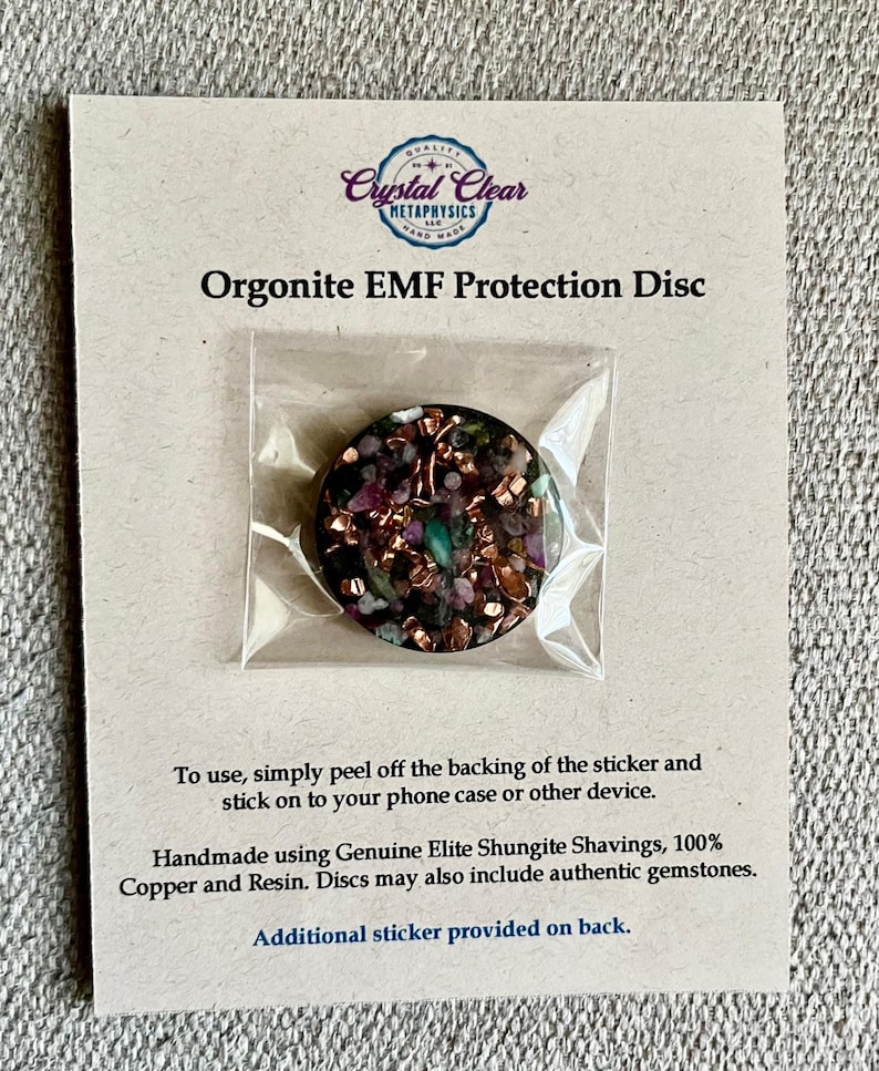May include: An Orgonite EMF Protection Disc, featuring a mix of copper, resin, and gemstones, is displayed in a clear plastic package. The disc is round with a dark, speckled appearance. The packaging includes text about the product's use and materials.