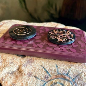 May include: Two round, black resin objects with copper accents rest on a rectangular, purple tray with a leopard print design. One object features a copper spiral, the other has copper and pink floral elements.