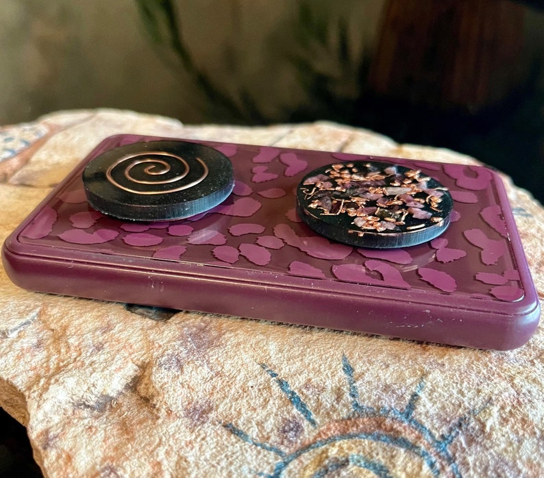 May include: Two round, black resin objects with copper accents rest on a rectangular, purple tray with a leopard print design. One object features a copper spiral, the other has copper and pink floral elements.