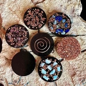 May include: A collection of seven black, circular orgone energy devices. Each device features a unique design, including copper wire, crystals, and a copper spiral. The crystals are various colors, including purple, blue, and light blue.