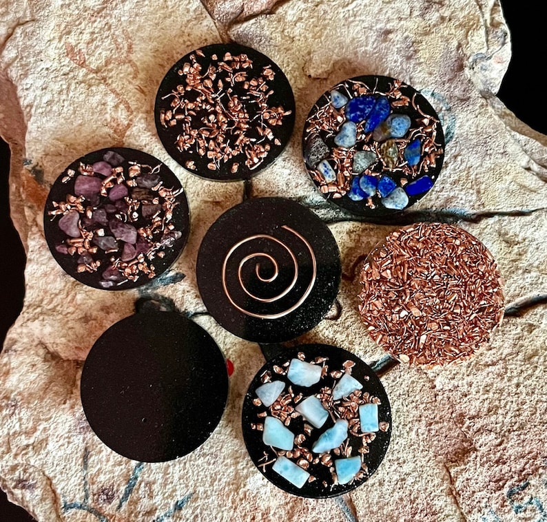 May include: A collection of seven black, circular orgone energy devices. Each device features a unique design, including copper wire, crystals, and a copper spiral. The crystals are various colors, including purple, blue, and light blue.