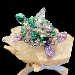May include: A crystal turtle sculpture with green, purple, and white crystals. The turtle is sitting on a clear quartz base.