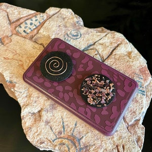 May include: A rectangular, purple trinket box with a leopard print design. Two black, circular objects are on top. One has a copper spiral, the other has small, purple and copper pieces. The box rests on a textured, tan stone.