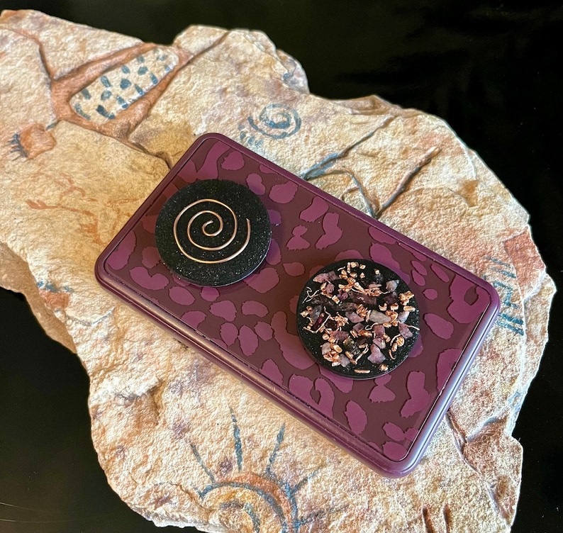 May include: A rectangular, purple trinket box with a leopard print design. Two black, circular objects are on top. One has a copper spiral, the other has small, purple and copper pieces. The box rests on a textured, tan stone.