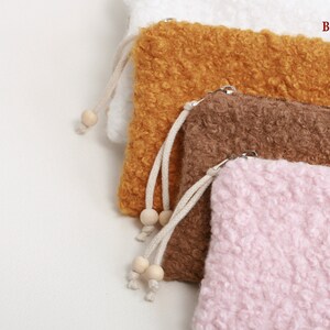 May include: Four fluffy, textured pouches in white, ochre, brown, and pink. Each pouch has a zipper closure and a drawstring with wooden beads. The pouches are labeled with the French color names: Blanc, Ocre, Marron, and Rose.