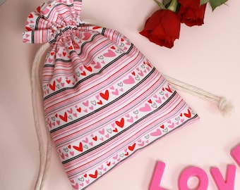 May include: A white drawstring bag with pink and red stripes and hearts. The bag is made of fabric and has a white drawstring. The bag is sitting on a pink surface with pink letters spelling out "LOVE" and a red heart.