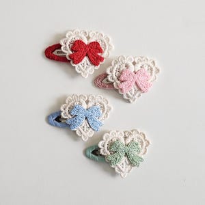 Pu&ograve; includere: Quattro fermagli per capelli a forma di cuore con dettagli in pizzo all'uncinetto e fiocchi. I fermagli presentano fiocchi rossi, rosa, blu e verdi, ciascuno su una forcina abbinata. Il pizzo &egrave; bianco, su sfondo bianco.