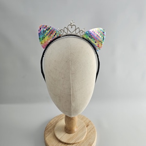 May include: A black headband with rainbow sequin cat ears and a silver crown. The headband is on a white mannequin head.