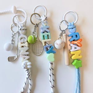 Op de afbeelding: Drie sportieve lanyards met witte koorden en witte plastic ringen. De eerste heeft een golfclub- en balbedel met kralen die GINA spellen. De tweede heeft een tennisracket- en balbedel met kralen die KELLY spellen. De derde heeft een honkbal- en knuppelbedel met kralen die RYAN spellen.