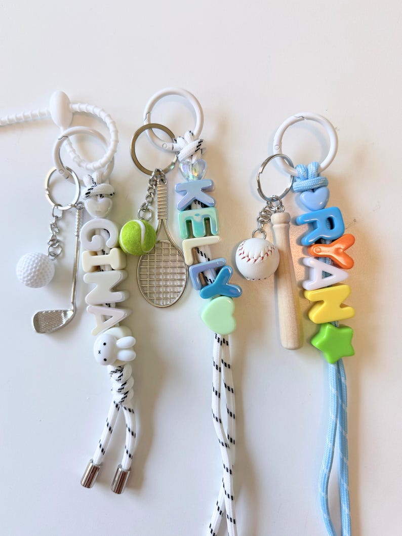 Op de afbeelding: Drie sportieve lanyards met witte koorden en witte plastic ringen. De eerste heeft een golfclub- en balbedel met kralen die GINA spellen. De tweede heeft een tennisracket- en balbedel met kralen die KELLY spellen. De derde heeft een honkbal- en knuppelbedel met kralen die RYAN spellen.