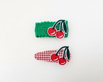Puede incluir: Dos pinzas para el pelo con bordado de cerezas rojas. Una pinza es verde y la otra es de cuadros rojos y blancos.